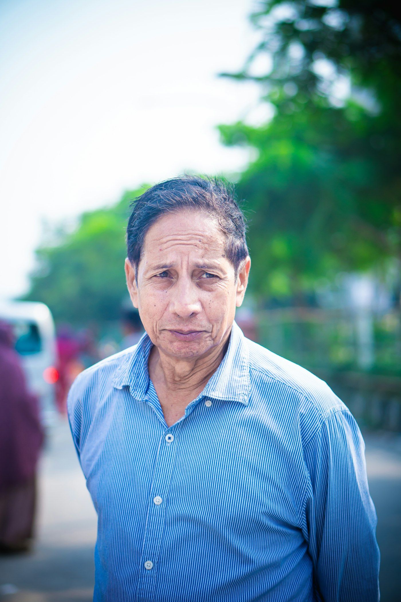 Man in a blue striped shirt photographed outdoors with green foliage behind him
