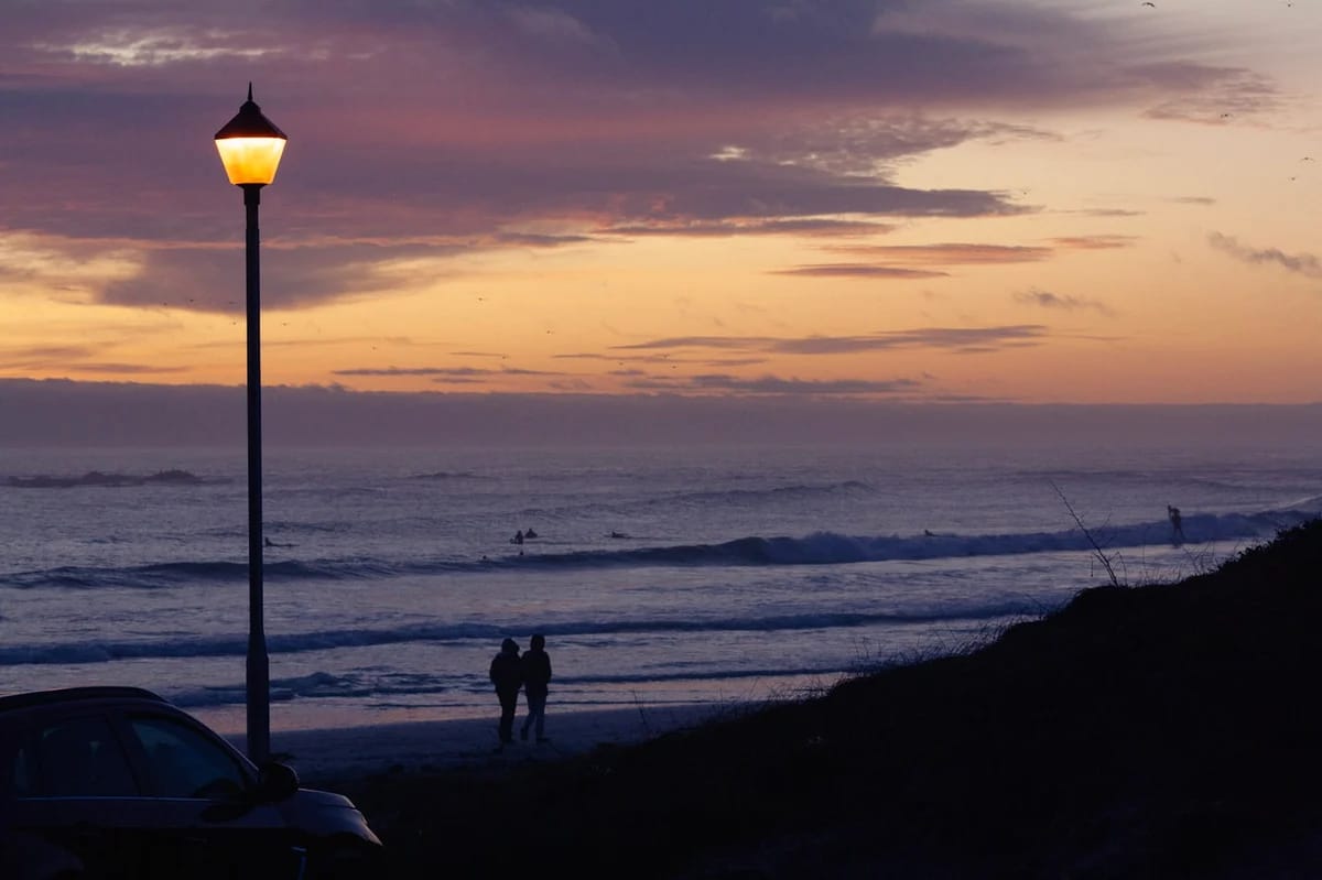 Couple standing on a beach cliff at dusk, silhouetted against a purple sky with surfers in the water below
