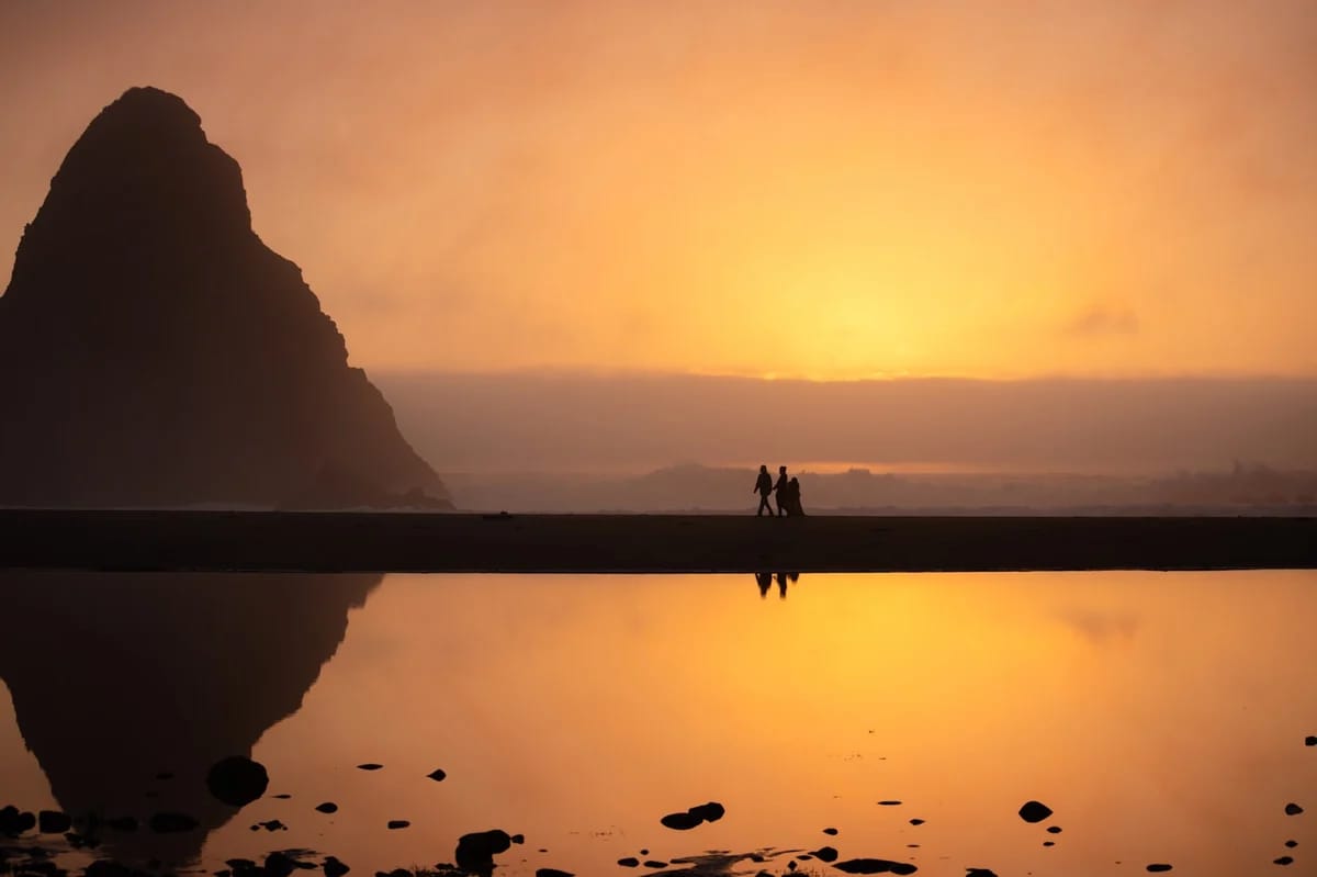 Silhouetted family walking along reflective beach at golden sunset with dramatic rocky cliff formation, calm waters, and glowing sky